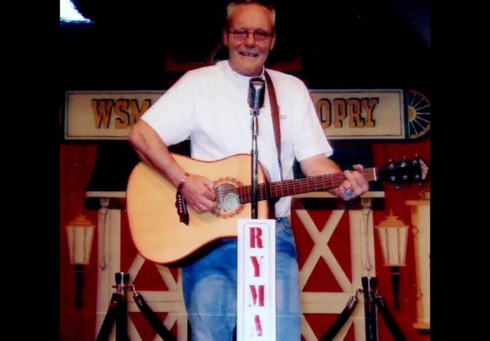 Big Gerry smiling at a microphone, playing an acoustic guitar